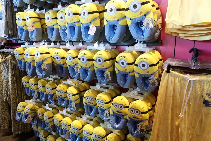 A retail wall display featuring numerous pairs of plush Minion-themed slippers in various sizes alongside matching yellow patterned pajama pants