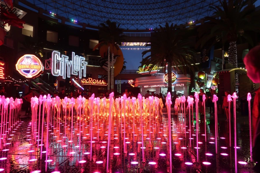 Vibrant pink illuminated dancing fountains at Universal CityWalk Hollywood at night, with glowing neon signs for Johnny Rockets and Bubba Gump Shrimp Co. under a large glass dome