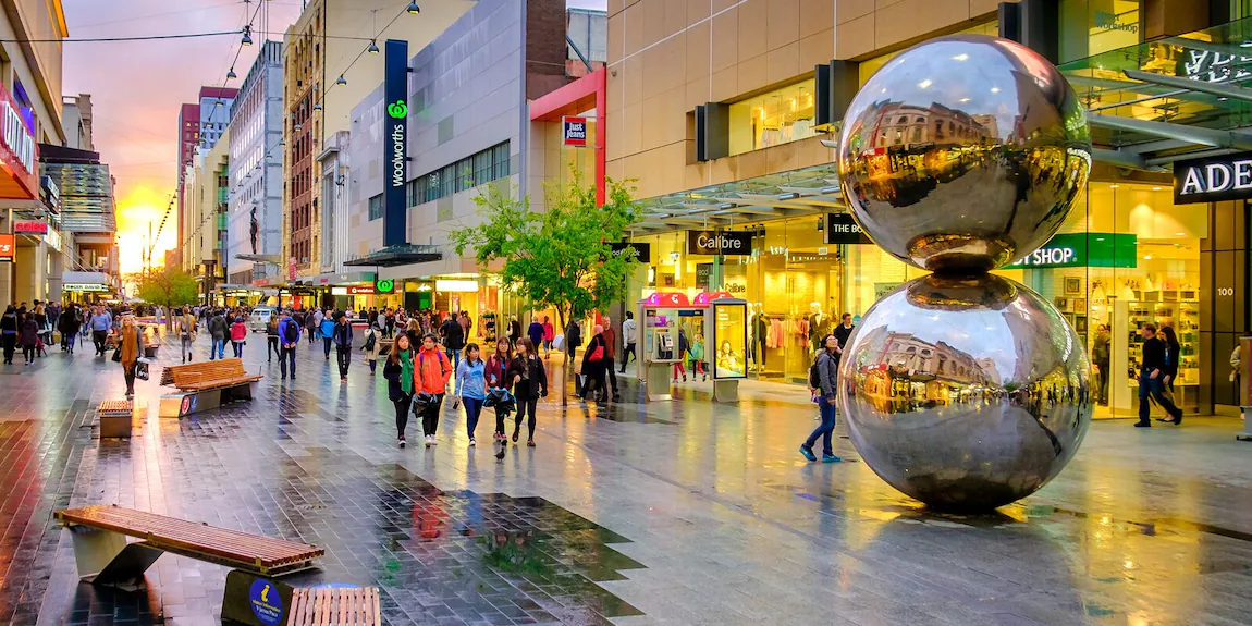 Shoppers walk along a wet pedestrian street next to the iconic shiny metallic Spheres sculpture outside storefronts at dusk.