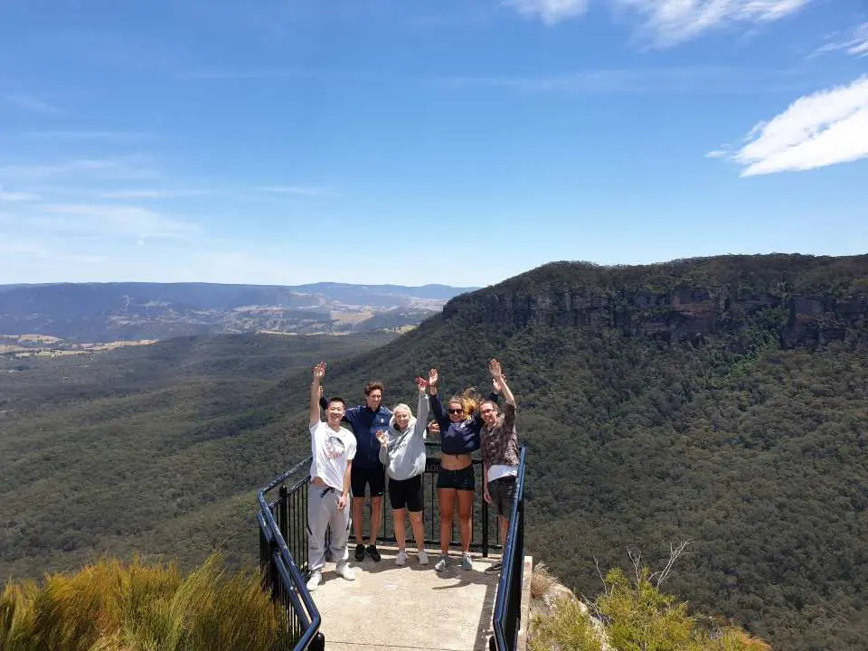 A group of five friends stands together on a fenced mountain lookout platform, waving their hands joyfully.