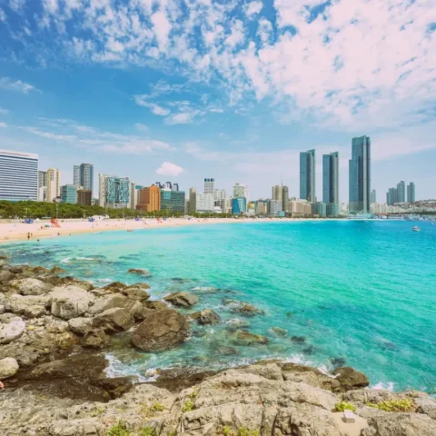 Panoramic view of Haeundae Beach in Busan featuring turquoise water, a rocky shoreline, and the city skyline under a blue sky.