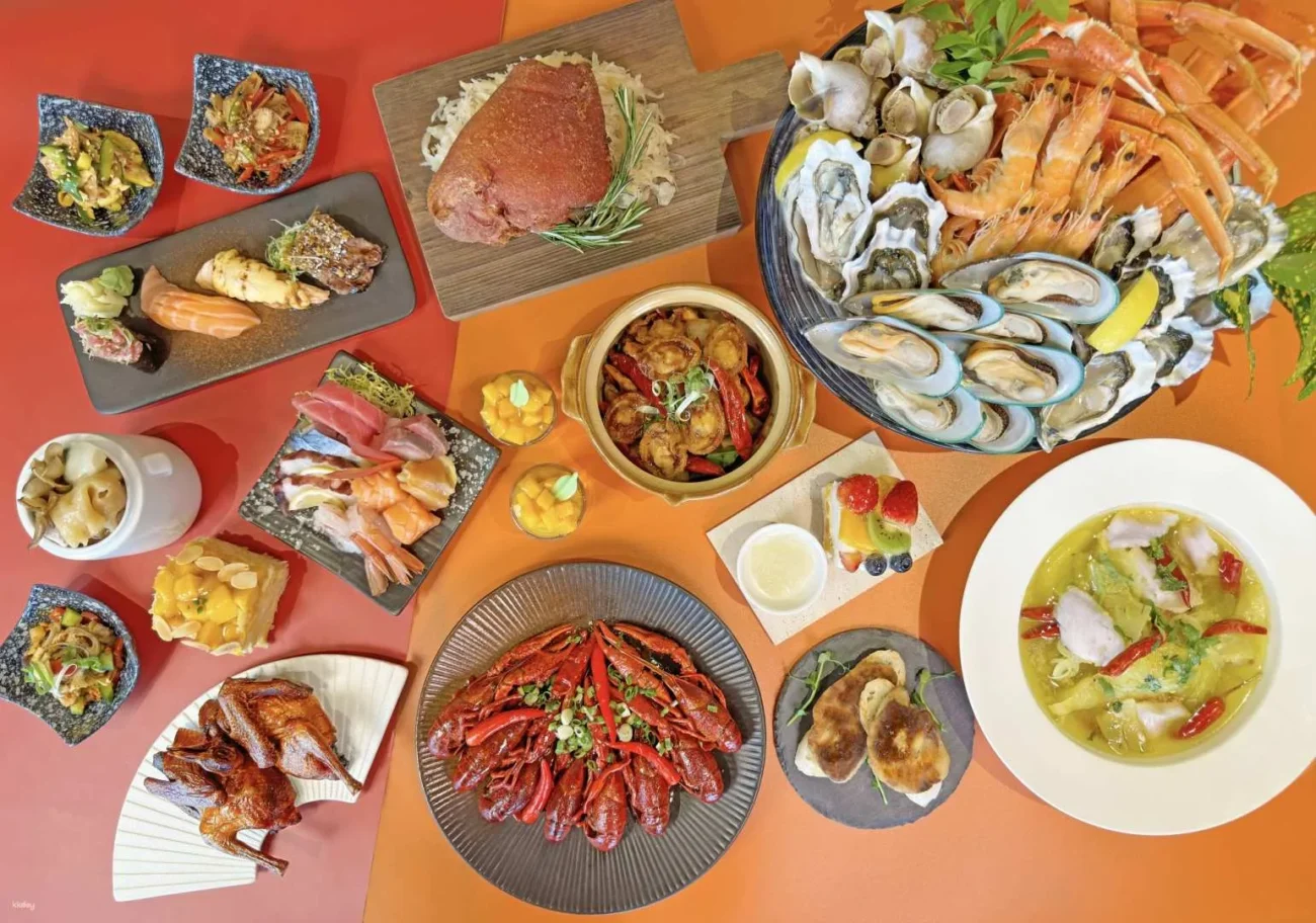 A large international buffet spread arranged on a dual-toned red and orange background. The variety includes a chilled seafood platter with crab legs, mussels, and oysters, alongside spicy crawfish, sashimi, and sushi.