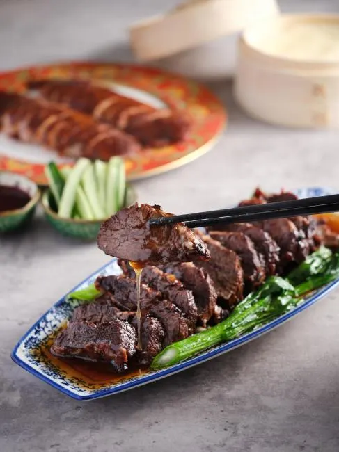 A pair of black chopsticks holds a succulent slice of braised beef brisket dripping with savory dark sauce. The meat is served on a blue and white patterned ceramic platter alongside vibrant green bok choy.
