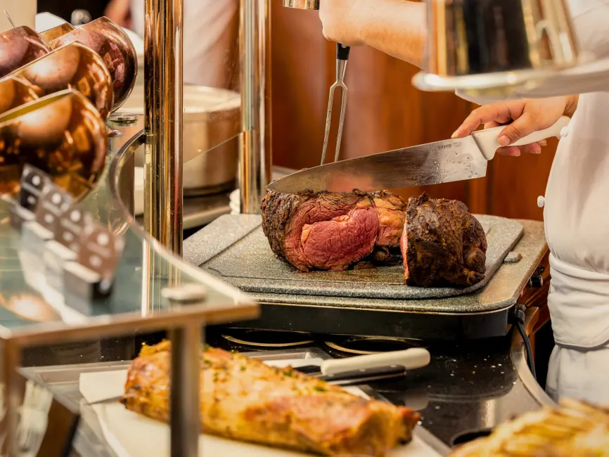 A chef in a white uniform carves a thick slice of medium-rare prime rib at a professional carving station. The roast beef has a dark, seasoned crust and a tender pink center.