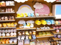 A brightly lit retail display shelf filled with various Disney Tsum Tsum plush toys, including characters like Winnie the Pooh, Donald Duck, and Mickey Mouse. A pink sign indicates a promotional price of 500 yen