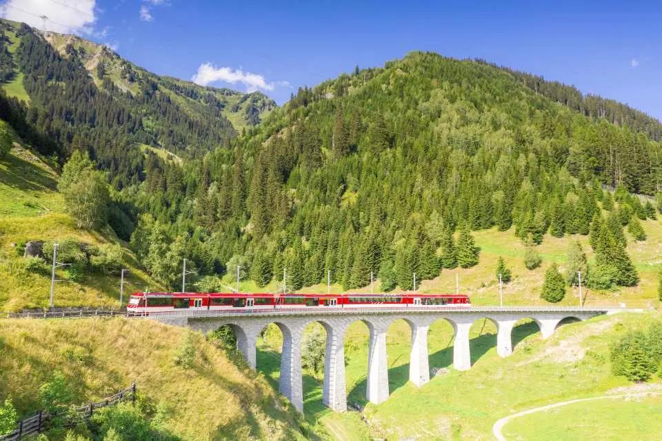Red train crossing a scenic stone viaduct through green mountains on a clear summer day in Europe