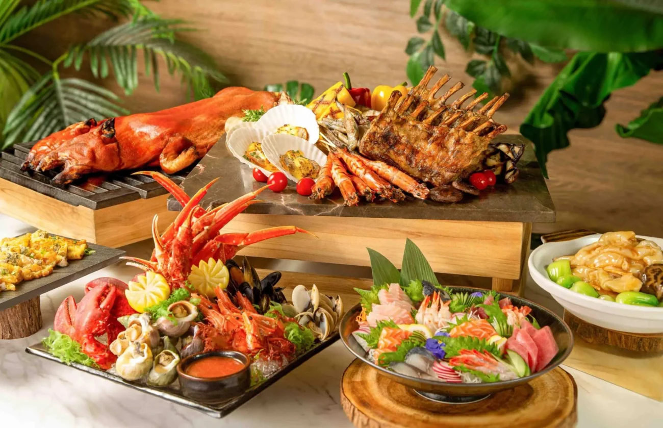 An elaborate gourmet buffet display featuring a roasted suckling pig, a rack of grilled lamb ribs, and various seafood platters. The spread includes a chilled seafood tower with lobster, crab legs, and prawns, as well as a large bowl of fresh sashimi including salmon and tuna.