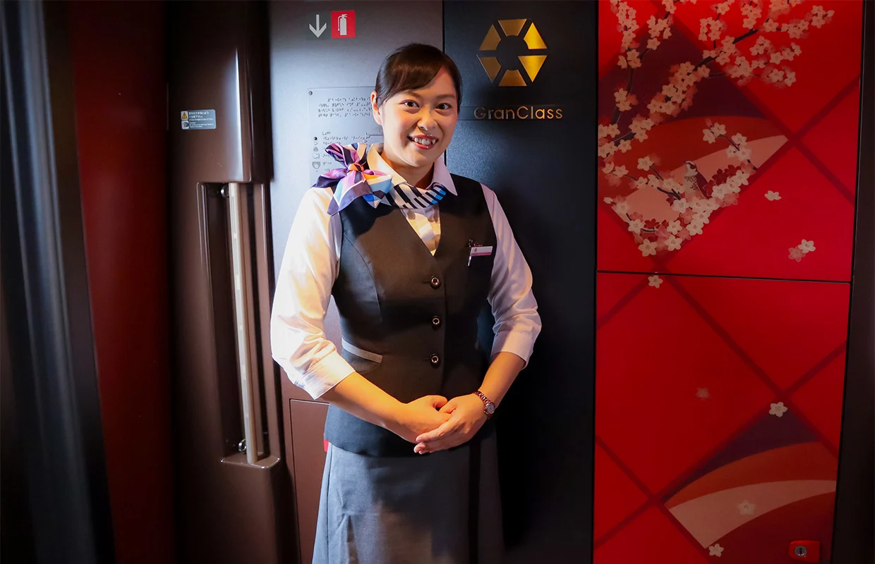 A friendly Gran Class Shinkansen attendant standing in the train cabin doorway, wearing a professional uniform, with the gold Gran Class logo visible on the dark wall.