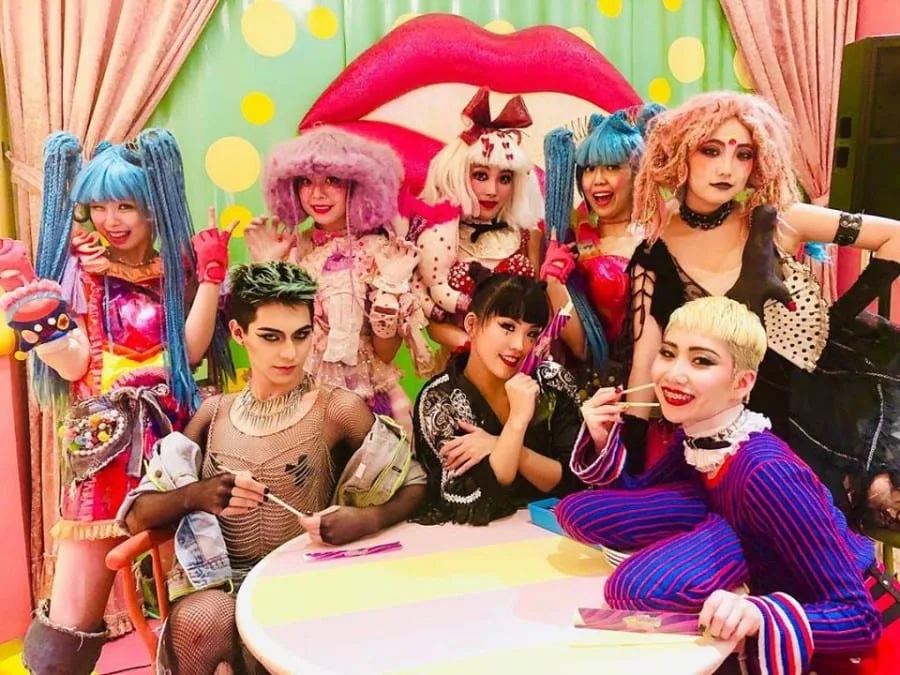 Group of performers dressed in colorful, eccentric Harajuku pop culture fashion posing around a table in Tokyo.
