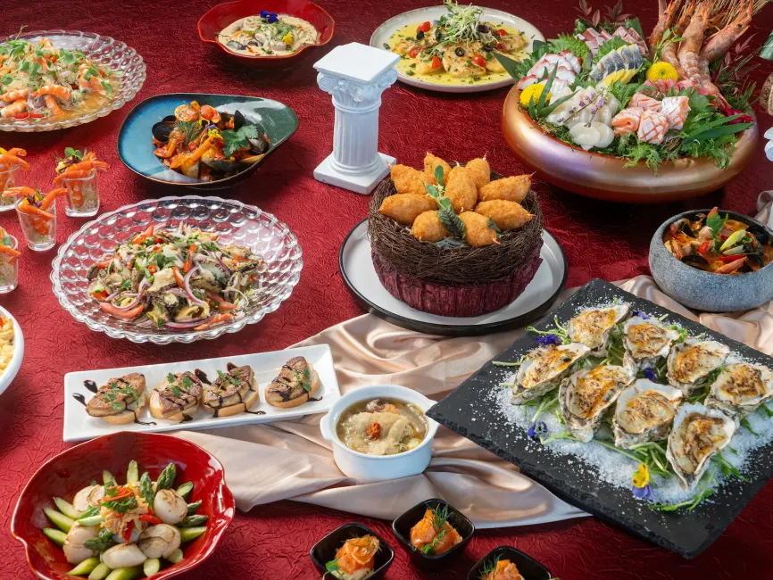 A luxurious international buffet spread displayed on a red textured tablecloth with a miniature white Greek column accent. The assortment includes a large sashimi platter with prawns and salmon, baked oysters on a bed of rock salt, foie gras crostini with balsamic glaze, and crispy fried crab claws served in a decorative nest.