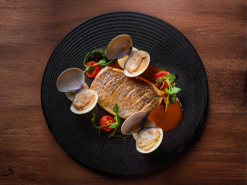 A top-down view of a gourmet seafood dish served on a textured black circular plate. The centerpiece is a pan-seared fish fillet with crispy golden-brown skin, surrounded by four steamed clams in their shells.