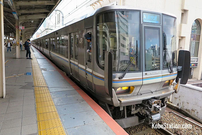 A silver JR West Special Rapid Service train stopped at Kobe Station platform in Japan, with a conductor looking out the cabin window.