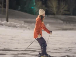 A person wearing a bright orange jacket, dark pants, and a beanie, skillfully skiing on a snowy slope during a winter evening.