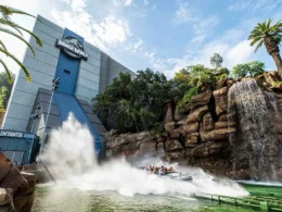 A theme park boat makes a large splash at the bottom of a steep drop on Jurassic World: The Ride, next to a rocky waterfall and a modern gray building at Universal Studios Hollywood