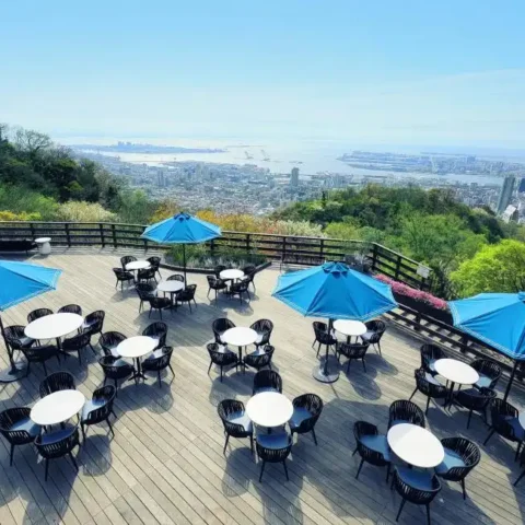 An expansive outdoor wooden terrace features neat rows of tables under bright blue umbrellas, showcasing a spectacular panoramic view of the city and harbor below.