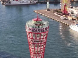 An aerial high-angle view of the top observation deck of the red Kobe Port Tower featuring the words PORT OF KOBE, overlooking the harbor and a nearby Ferris wheel.