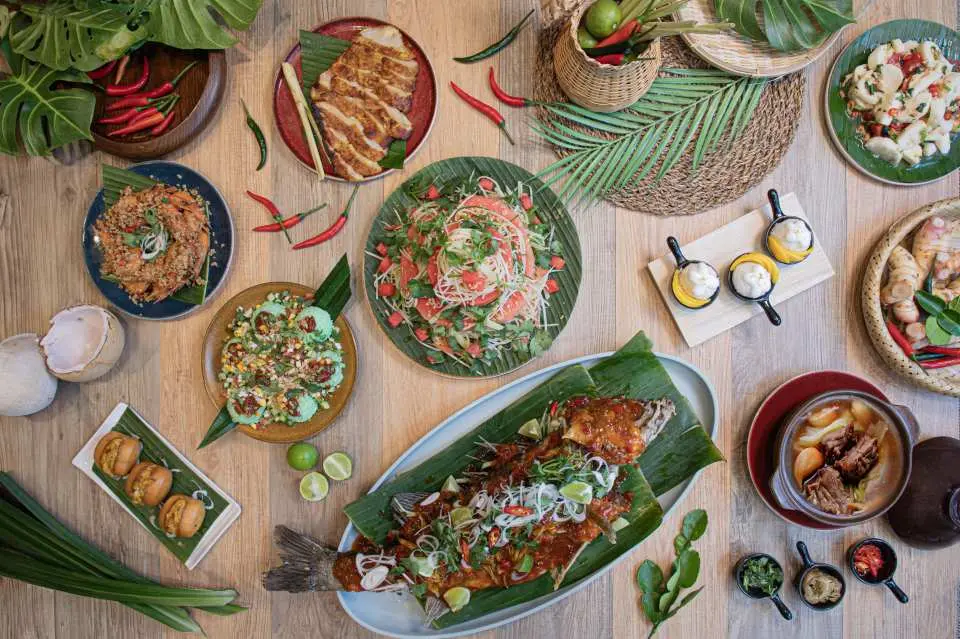 A top-down view of a traditional Southeast Asian feast spread across a light wooden table. The centerpiece is a large whole steamed fish topped with chili sauce, herbs, and lime on banana leaves.
