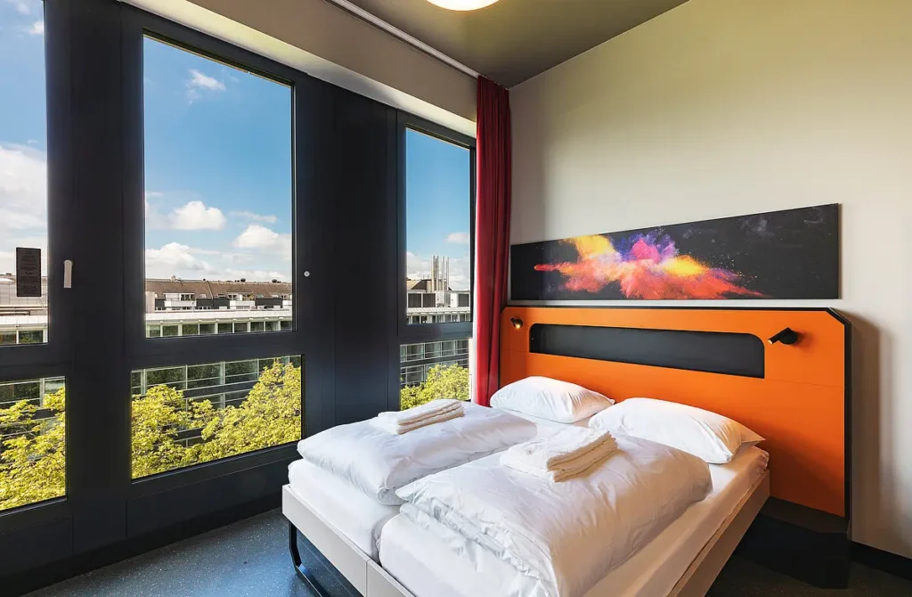 A modern double hotel room featuring an orange headboard, white duvets, and large floor-to-ceiling windows showing a bright city view with green trees.