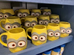 A collection of bright yellow ceramic mugs shaped like Minion characters with large goggle eyes and small smiles, arranged on a blue wooden shelf