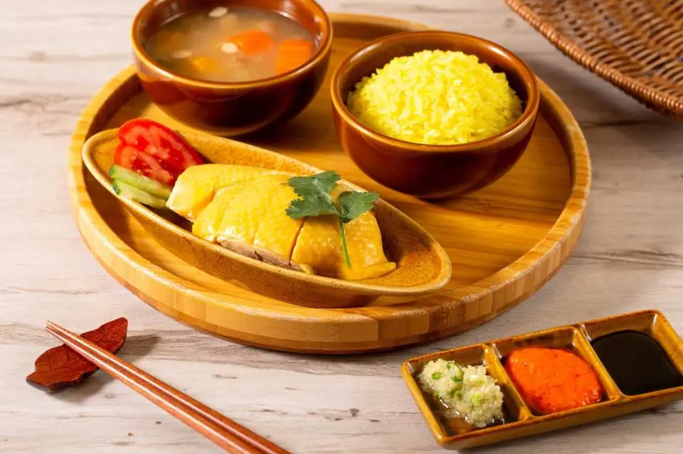 A traditional Hainanese chicken rice set served on a round wooden tray. The meal includes a plate of tender, sliced poached chicken with yellow skin, a bowl of fragrant yellow ginger rice, and a bowl of clear vegetable soup with carrots.