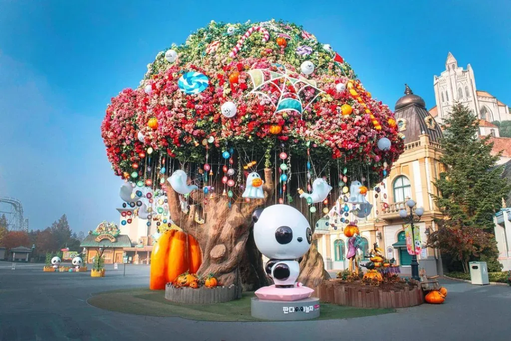 A large artificial tree decorated with colorful flowers, lollipops, ghost figurines, and giant pumpkins, featuring a panda statue at its base in Everland Theme Park.