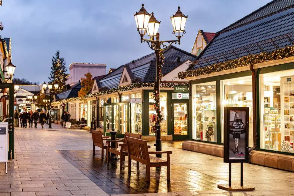 A European-style outdoor shopping village at dusk decorated with warm holiday lights, featuring store displays, street lamps, and wooden benches along a paved walkway.