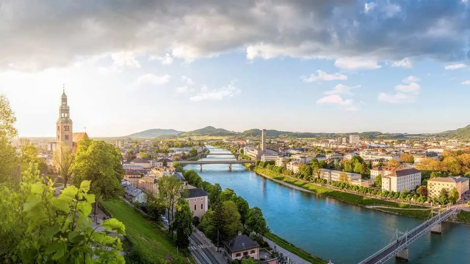 High-angle panoramic view of Salzburg city showing the Salzach River flowing between historic buildings, bridges, and green trees under a cloudy sky.