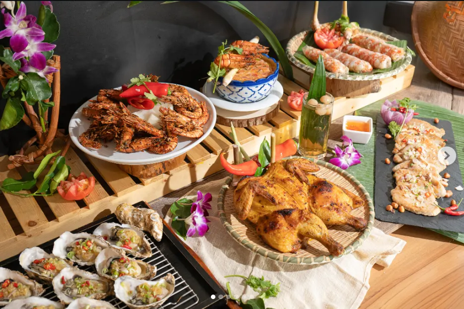 A vibrant spread of Vietnamese and Thai fusion dishes displayed on wooden slats and banana leaves. The table includes grilled whole chicken, garlic oysters on a half shell, crispy salt and pepper prawns, fresh spring rolls, a spicy coconut soup, and grilled pork slices.