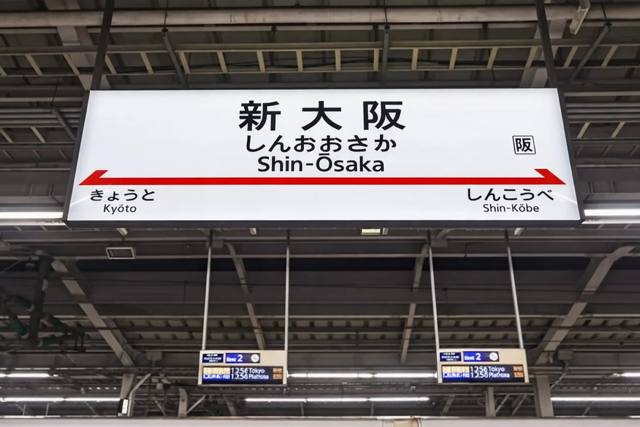 A hanging Japanese train station sign for Shin-Osaka Station. The sign displays the name in Kanji, Hiragana, and English Romaji, with arrows pointing toward the adjacent stations, Kyoto and Shin-Kobe.