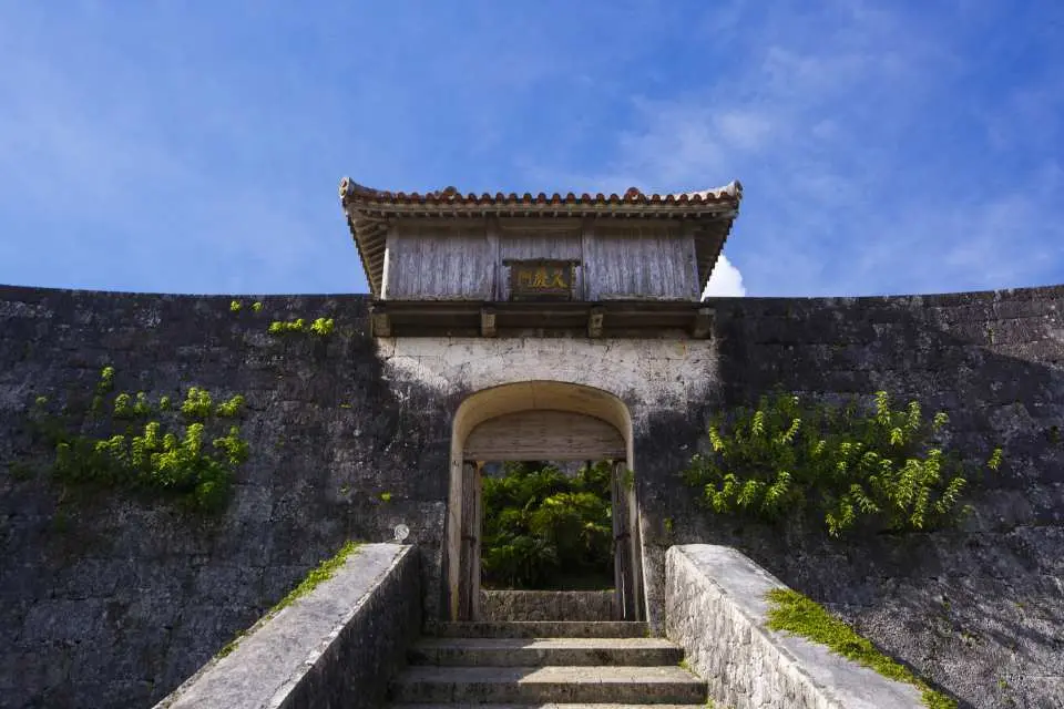 The historic stone fortress wall and traditional wooden gate of Shurijo Castle in Okinawa under a bright blue sky.