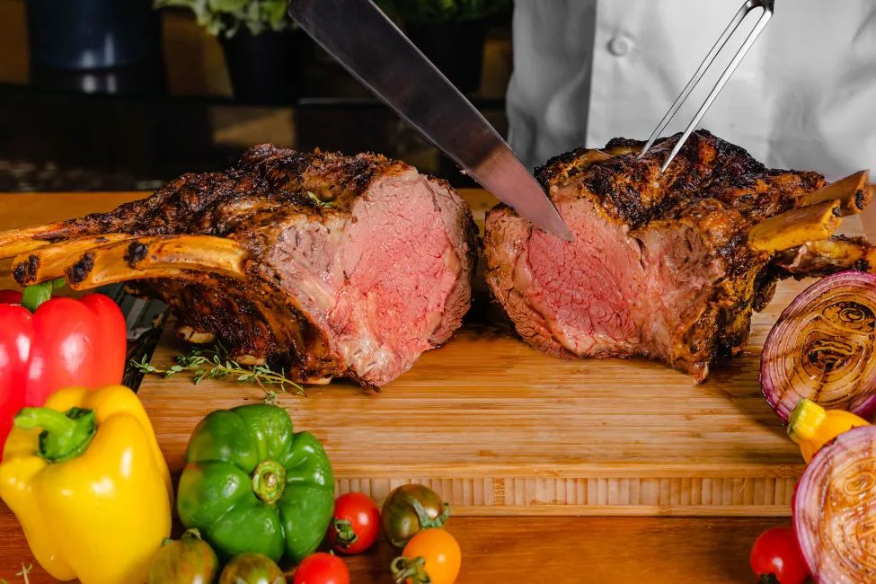 A chef in a white uniform carves a large, herb-crusted bone-in ribeye roast on a wooden cutting board. The meat is cooked to a perfect medium-rare with a deep brown sear and a juicy pink interior.