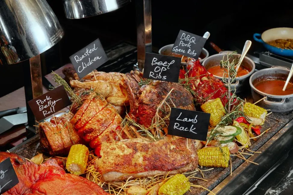 A gourmet roasted meat station at a professional buffet featuring several large cuts of meat under heat lamps. The selection includes a crispy pork belly roll, a whole roasted chicken, beef OP ribs, and a lamb rack, each marked with a chalkboard label.