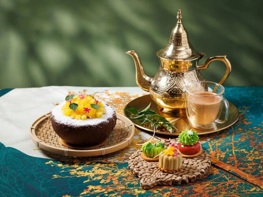 An elegant Middle Eastern dessert spread featuring a chocolate bowl filled with coconut shavings and diced mango. Next to it are three colorful petit fours shaped like cacti and flowers resting on a woven coaster.