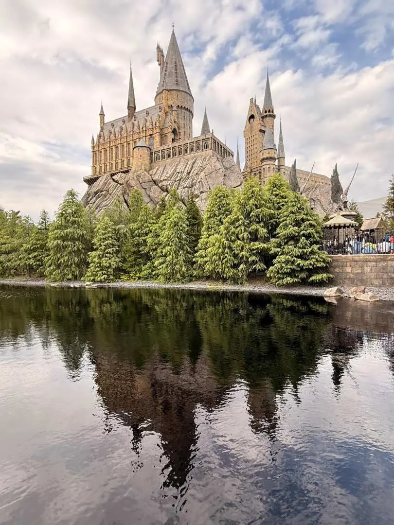 Hogwarts Castle standing on a rocky cliff, perfectly reflected in the still waters of the Black Lake at Universal Studios Japan under a cloudy sky.