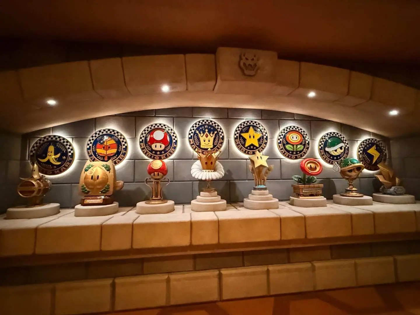 A row of eight golden Mario Kart trophies, including the Mushroom, Flower, and Star Cups, displayed on a tiered stone ledge under warm lighting inside the Bowser's Castle queue at Super Nintendo World, Universal Studios Japan.