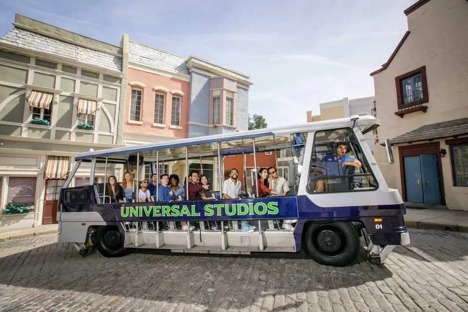 A white and blue Universal Studios tour tram filled with passengers driving through a European-style movie set street with colorful buildings