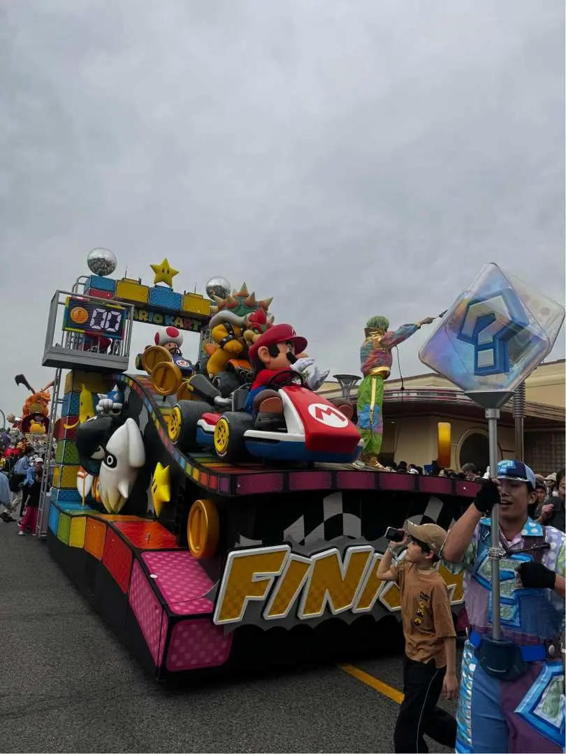 A vibrant Mario Kart parade float at Universal Studios Japan featuring Mario, Bowser, and Toad on a rainbow road track with a "FINISH" sign and floating question blocks.