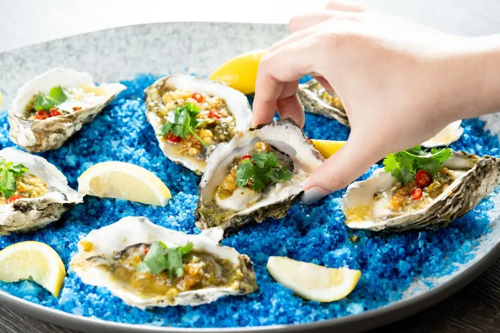 A hand with grey painted fingernails is reaching for a raw oyster on a half shell served in a large grey platter. Multiple oysters in half shells, topped with minced garlic, chili peppers, and fresh cilantro leaves, are arranged on a bed of vivid blue rock salt.