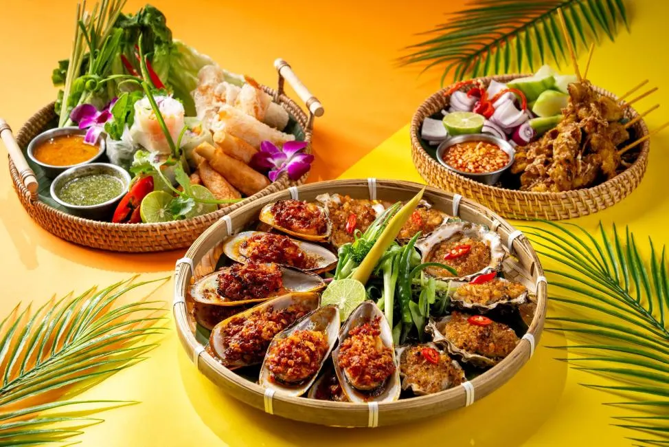 A colorful Southeast Asian appetizer spread presented in woven bamboo baskets on a bright yellow background. The central basket features steamed mussels and oysters topped with spicy minced garlic and chili. Other baskets include fresh spring rolls with dipping sauces and grilled chicken satay skewers served with a peanut sauce, red onions, and cucumbers.