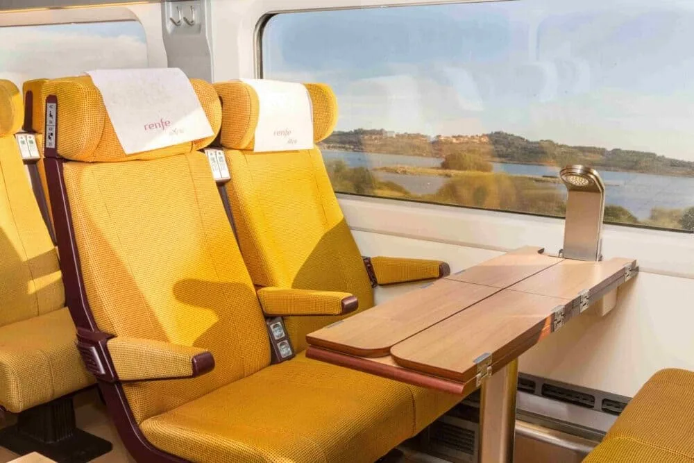 Interior of a Renfe train featuring bright yellow upholstered seats with a wooden folding table and a large window overlooking a lake.