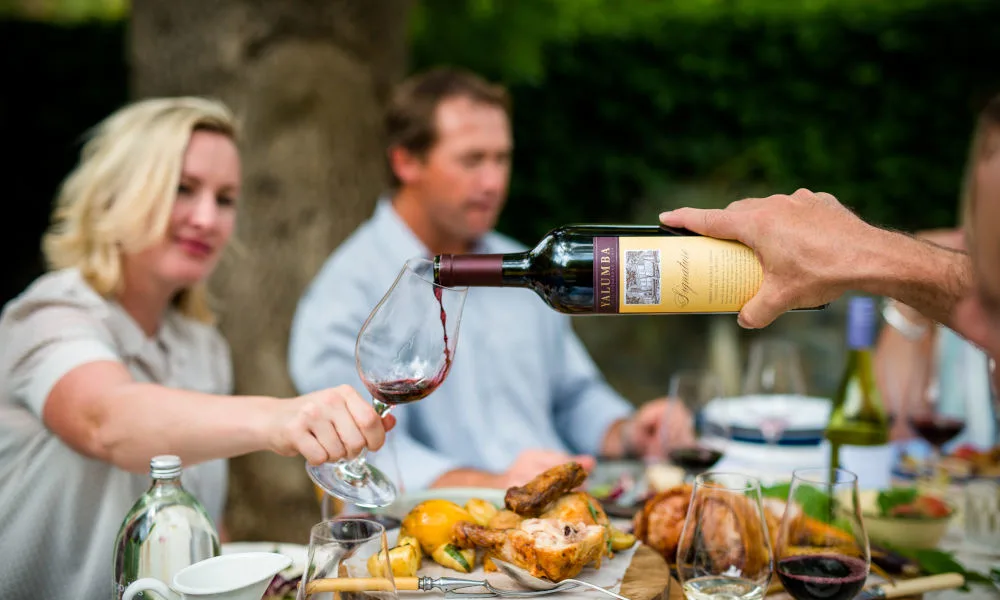 A hand pours red wine from a Yalumba bottle into a glass held by a woman during an outdoor dinner party with friends.