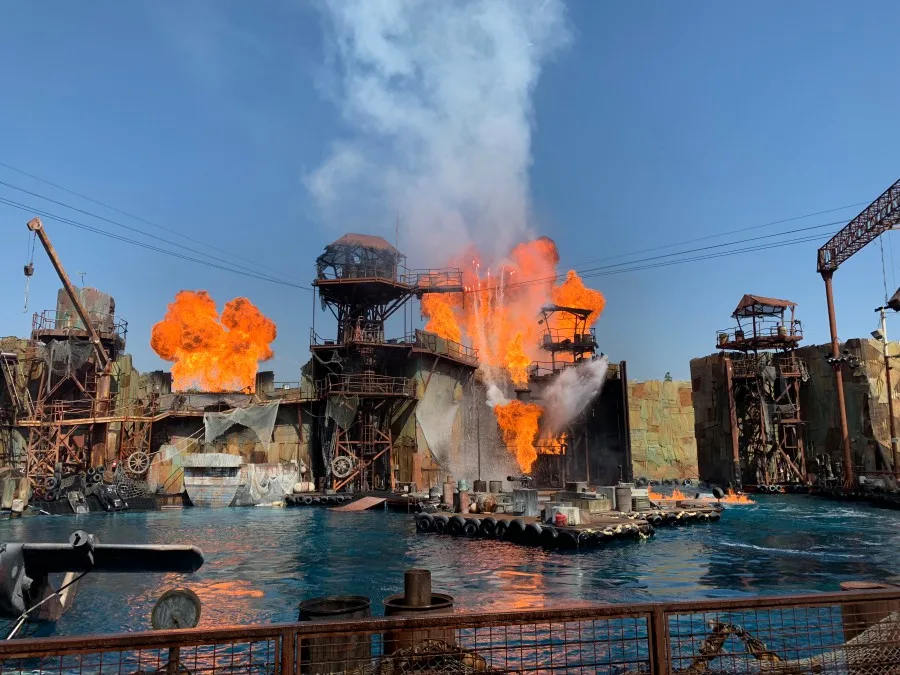 A live performance of the WaterWorld stunt show at Universal Studios, featuring massive orange fire explosions, white smoke plumes, and water splashes