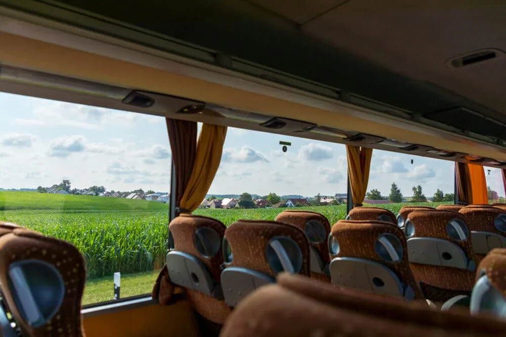View from inside a bus showing brown patterned seats and large windows overlooking a green field and rural village.