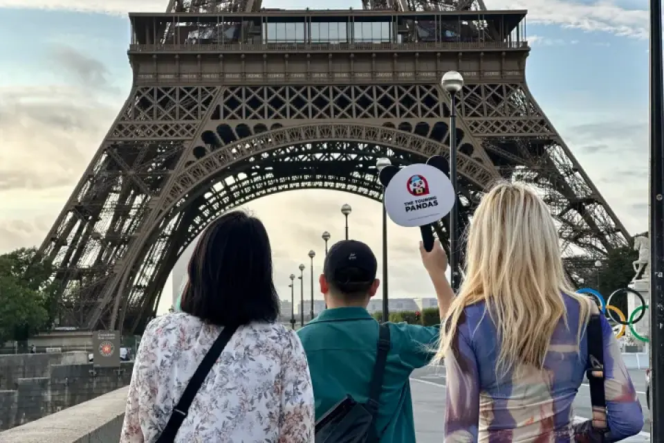 Rear view of three travelers standing before the Eiffel Tower in Paris, with one holding a "The Touring Pandas" sign near the Olympic rings.