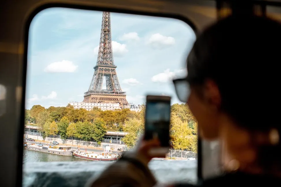View through a train window of a person taking a smartphone photo of the Eiffel Tower in Paris, France, overlooking the Seine River.