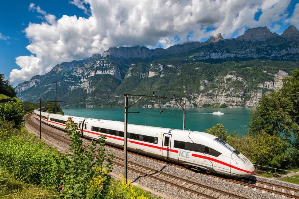A white and red high-speed ICE train traveling along a scenic railway next to a turquoise lake and massive rocky mountains in Switzerland.
