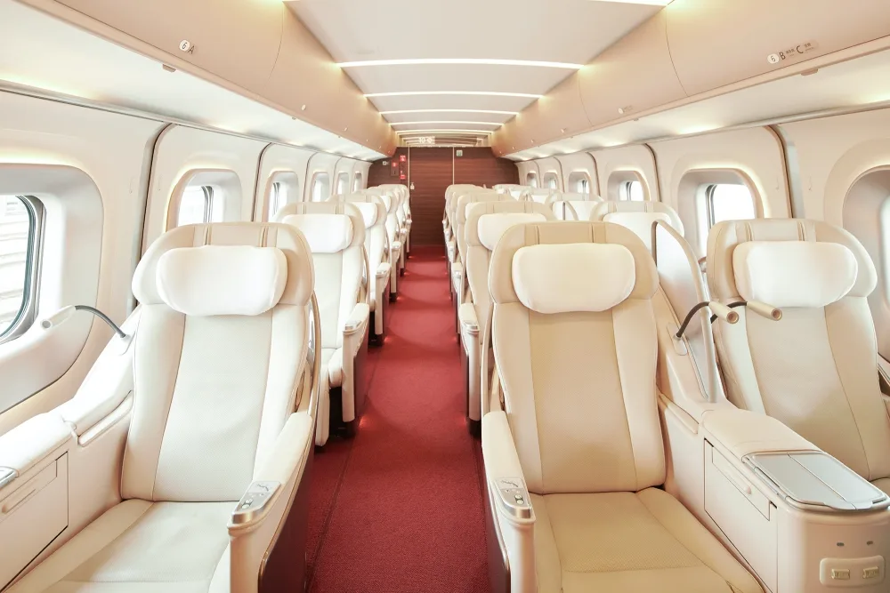 Spacious and modern interior of a Gran Class Shinkansen train cabin featuring cream-colored leather reclining seats in a 1+2 layout on a red carpet.