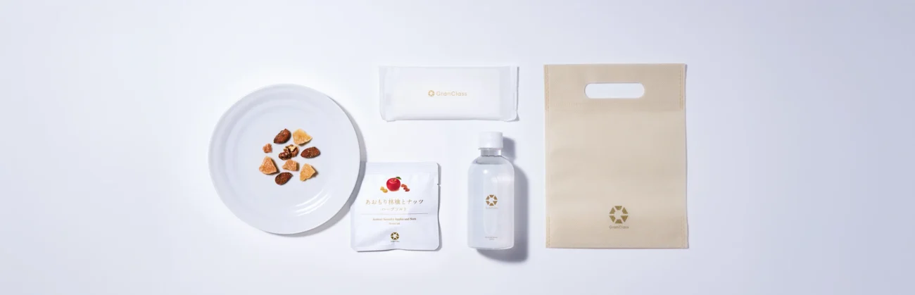An overhead view of a Gran Class Shinkansen welcome amenity kit, including a bottle of water, dried fruit and nut snacks, a wet towel, and a branded pouch on a white background.
