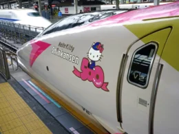A close-up of the Hello Kitty Shinkansen 500 series high-speed train operated by JR-West at a station platform in Japan, featuring a white, pink, and lime green livery with the Hello Kitty characte