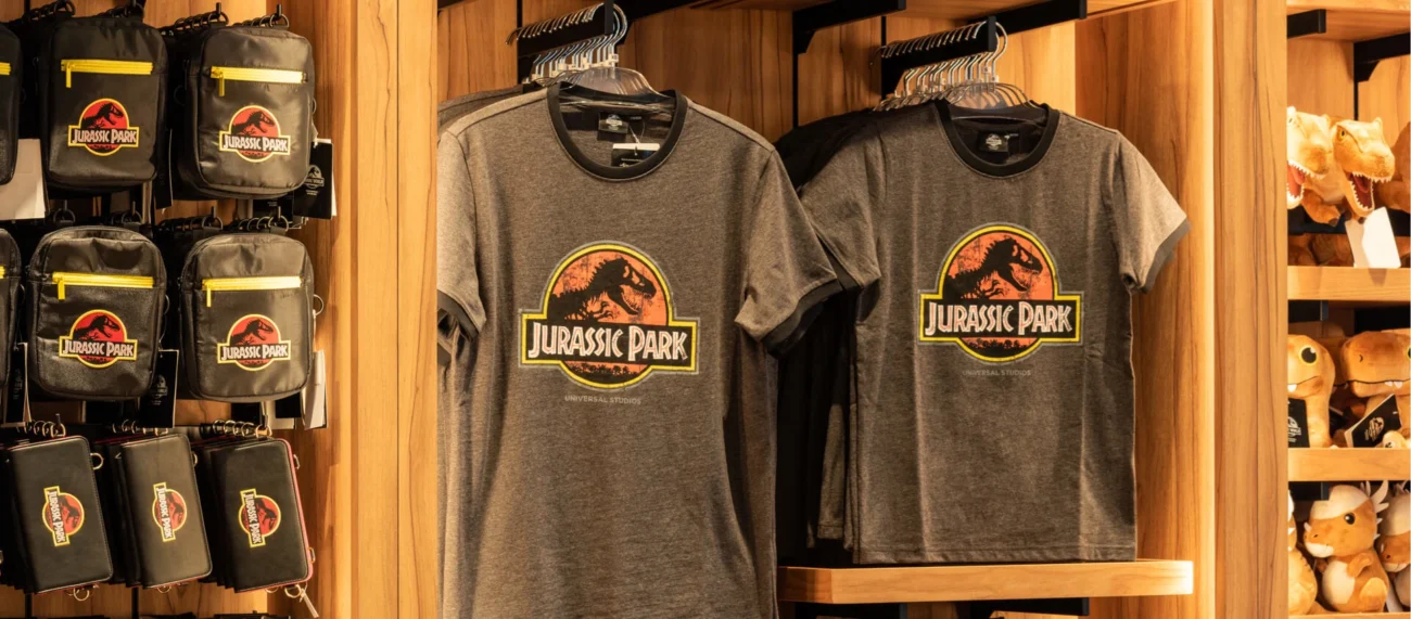 Retail display at Universal Studios showing gray Jurassic Park t-shirts, black crossbody bags, and dinosaur plush toys on wooden shelves.